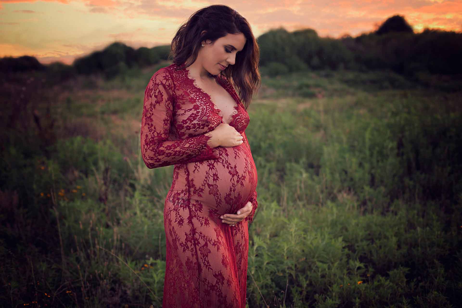 A pregant woman in a red see through robe standing in an open field during sunset